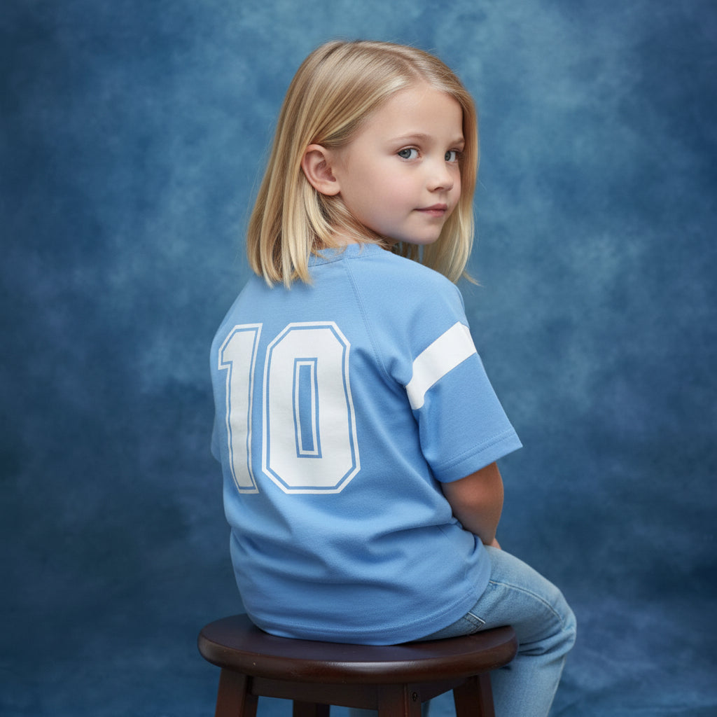 Young girl wearing a Blue Jogiel Blue 10 Raglan shirt with '10' printed on the back, sitting on a stool against a blue yearbook background.