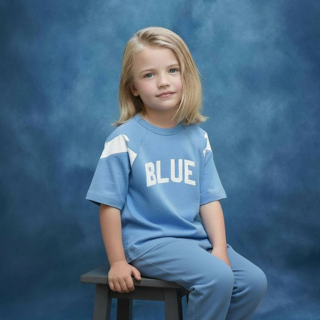 Young girl wearing a Blue Jogiel Blue 10 Raglan shirt with 'BLUE' printed on it, sitting on a stool against a blue yearbook background.