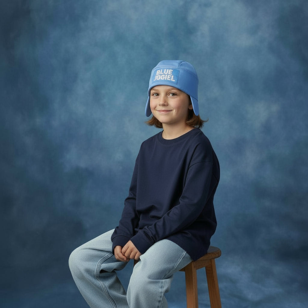 Child wearing a Blue Jogiel Ear Flap Hat and navy sweater sitting on a wooden stool against a blue yearbook background