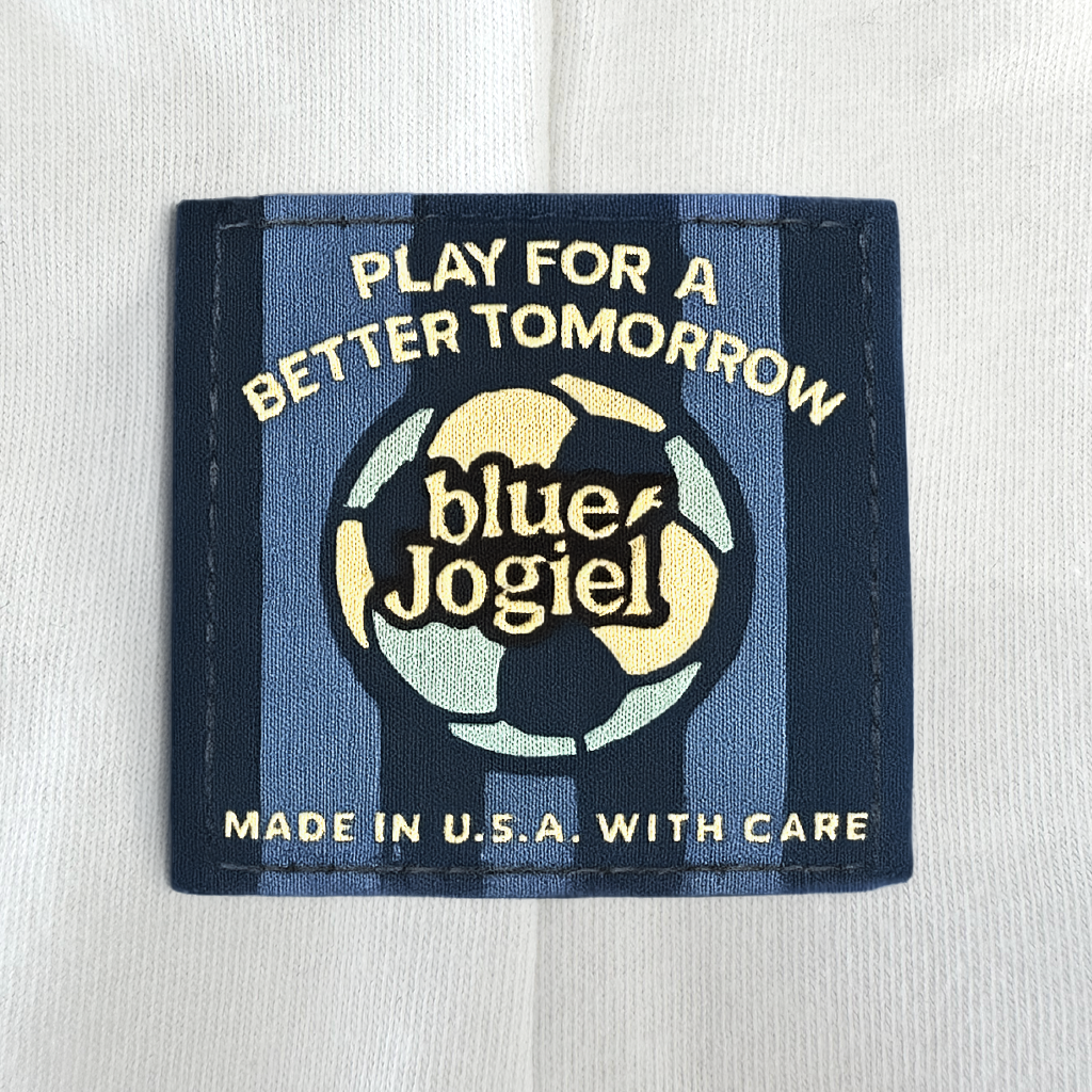 Woven label with 'Play for a Better Tomorrow' text and 'Blue Jogiel' logo and soccer ball sewn onto a white garment on the seam.