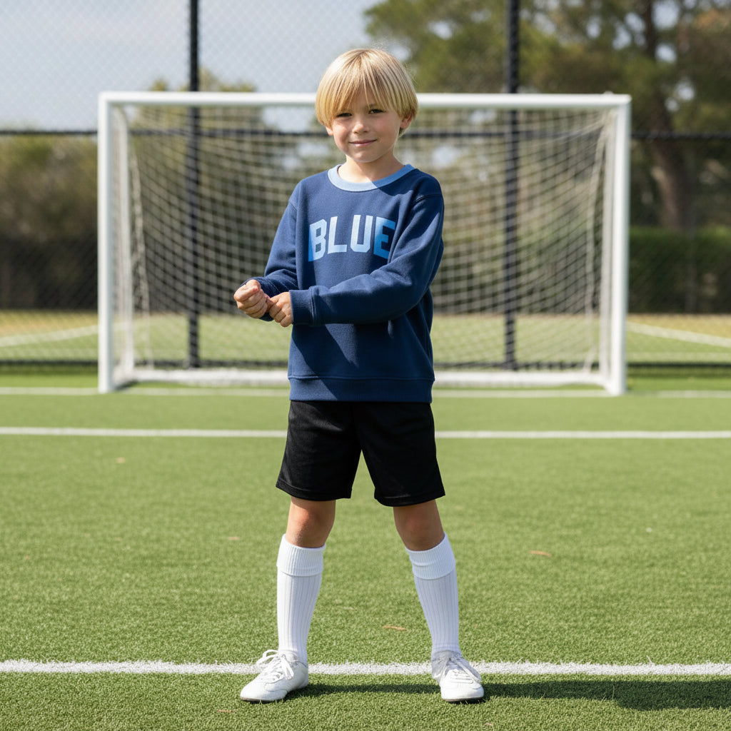 Young boy wearing a navy blue Blue Jogiel 'Soccer Academy Crewneck' sweatshirt standing on soccer field in front of a soccer goal.